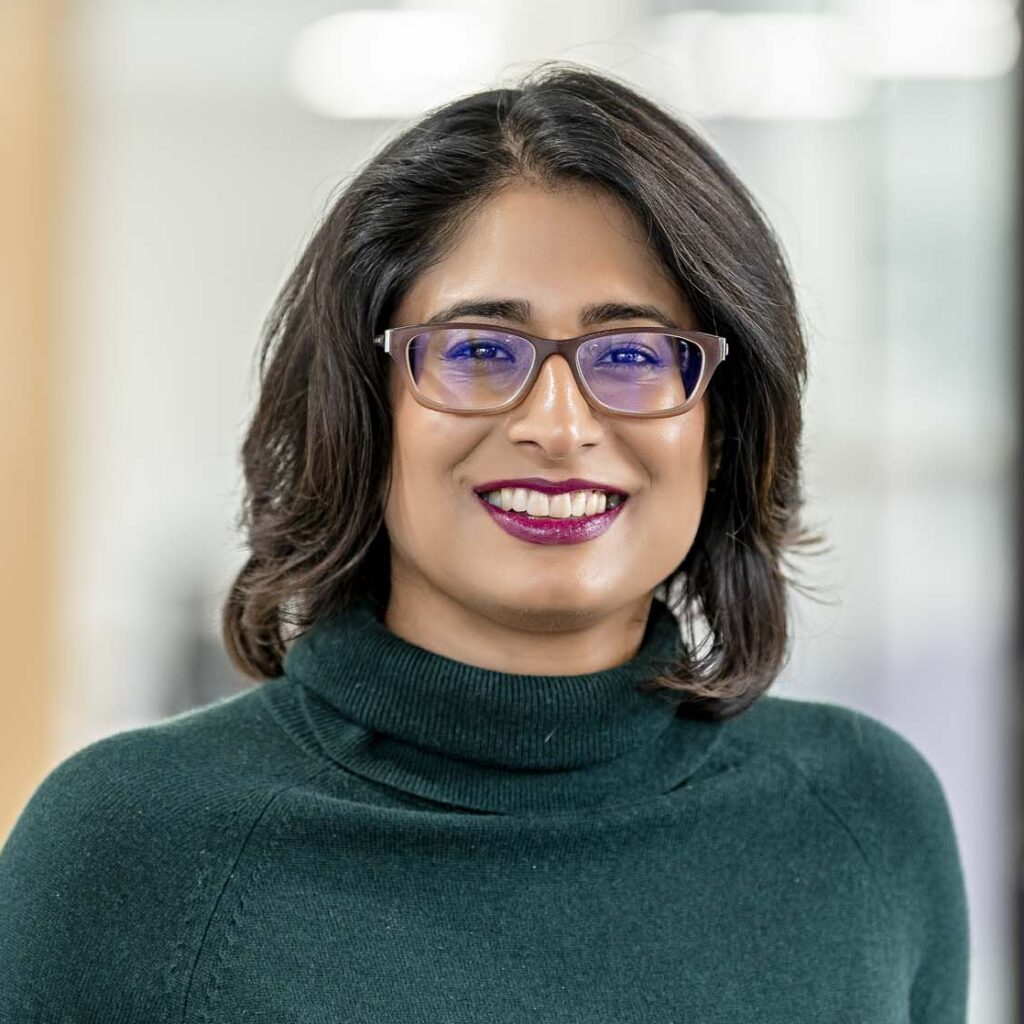 A Person With Shoulder-Length Dark Hair And Glasses, Reminiscent Of A Finance Executive, Smiles While Wearing A Green Turtleneck. The Background Is Blurred.