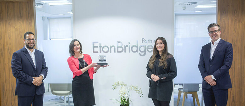 Four People Stand In An Office With An &Quot;Etonbridge&Quot; Sign; One Woman Proudly Holds A Plaque Reading &Quot;100.&Quot; Eton Bridge Partners Placed Number 14 In The Uk'S 100 Best Small Companies To Work For List, Celebrating Their Outstanding Workplace Culture And Commitment To Excellence.