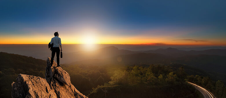 A Person In Business Attire Stands On A Rocky Peak, Gazing At A Sunset Over A Mountain Landscape With A Winding Road Below, Contemplating Insights From The Institute Of Interim Management Survey 2021.