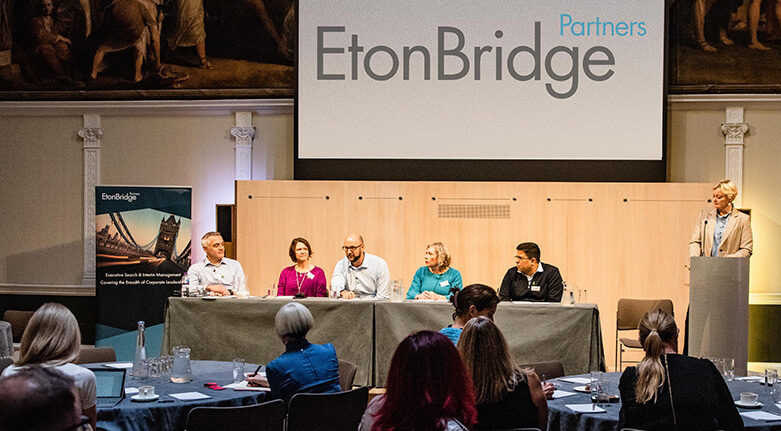 A Panel Of Six People Speaks At Eton Bridge Partners’ 5Th Annual Wellbeing Event 2021, With A Large Logo Displayed On A Screen Behind Them And An Audience Seated In Front.