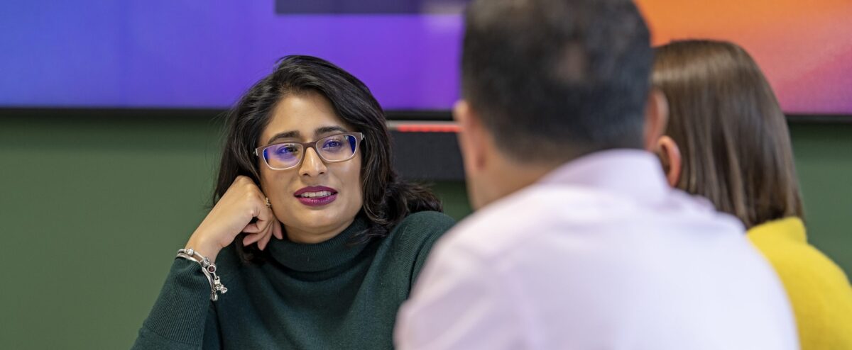 Three people sitting at a table in discussion, with one woman in glasses listening attentively, fostering an atmosphere of inclusion.