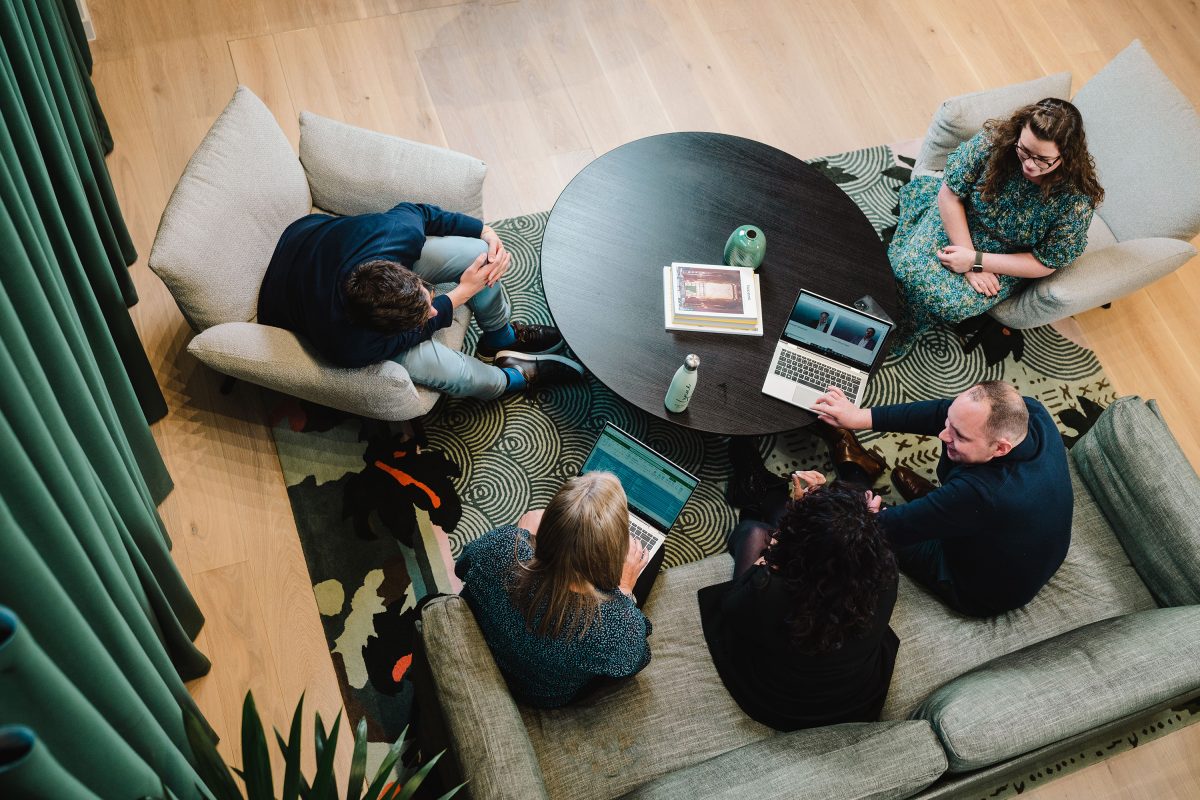 Diverse group from EBP team, birdseye view, sitting around a table on sofas, reviewing a piece of work over a laptop